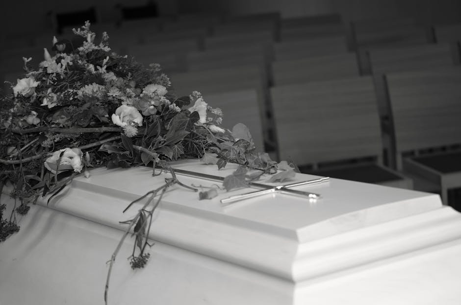 A funeral casket covered with a large, lush floral arrangement consisting of white and pale-colored flowers, such as roses and lilies, intertwined with green foliage. The arrangement is placed on top 