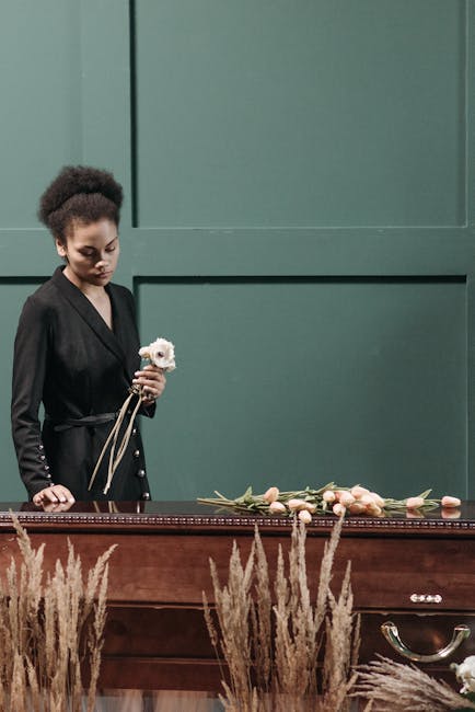 A young woman with dark curly hair, dressed in a black blazer, holding a small white flower with pink accents in her left hand, stands beside a wooden casket adorned with a floral arrangement of pale 