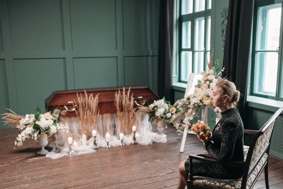 A woman seated on an elegant patterned armchair inside a softly lit room with green-paneled walls and large windows, holding a bouquet of orange flowers. In front of her, a floral arrangement featurin