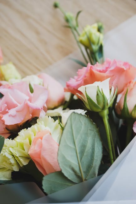 A close-up of a floral arrangement featuring pink and cream lisianthus flowers with ruffled petals, complemented by fresh green leaves and stems. The bouquet is wrapped in white paper and appears to b