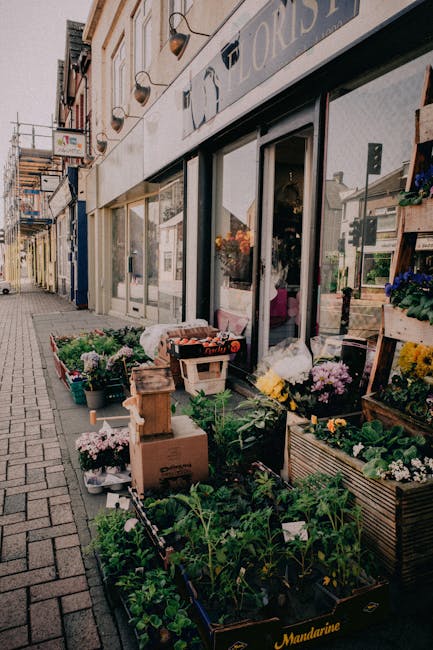 An outdoor display of fresh flowers and potted plants arranged along the sidewalk outside a florist shop, featuring colorful blooms including pink chrysanthemums, yellow daisies, purple petunias, and 