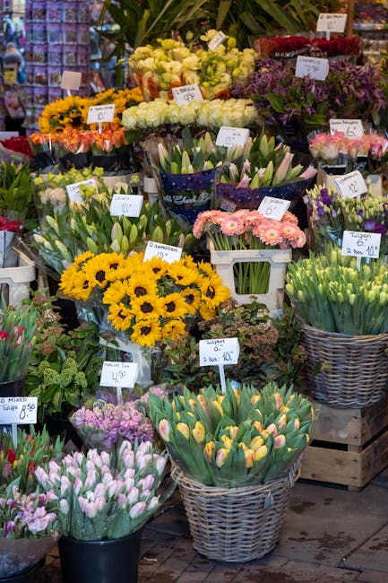 A vibrant display of fresh flowers at a florist shop, featuring bouquets of yellow sunflowers, white roses, and pink tulips arranged in baskets and wrapped bouquets. The flowers are neatly organized o