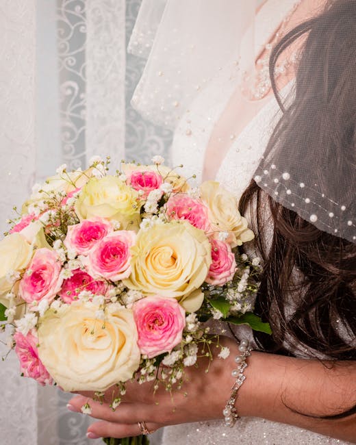 A bride holding a bouquet of fresh flowers featuring large ivory roses and smaller pink roses, accented with delicate white baby's breath and green foliage. The bouquet is wrapped with a simple ribbon