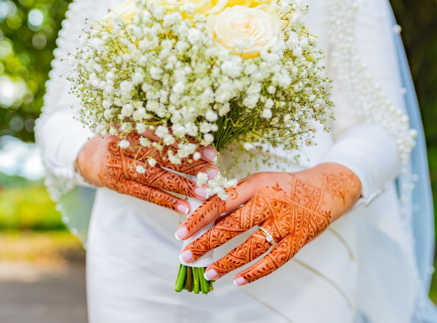 A person dressed in white, holding a bouquet of fresh flowers featuring white roses and baby's breath, wrapped in white paper and tied with a white ribbon. The individual's hands display intricate hen