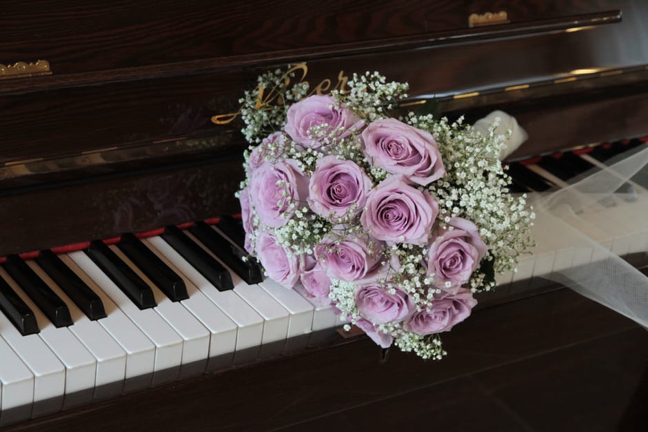 A bouquet of fresh, pale lavender roses arranged with delicate white baby's breath rests on the black and white keys of a polished wooden piano, near the gold-embossed nameplate. The roses are evenly 